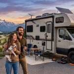 A happy couple with their small dog standing beside a Class C camper van at a scenic mountain overlook during sunset, equipped with solar panels and a Starlink satellite dish on the roof.