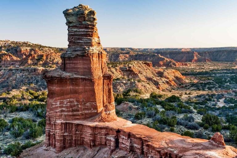 Tall, layered red rock formation standing prominently in a vast desert canyon landscape under a clear blue sky. best RV parks in Texas